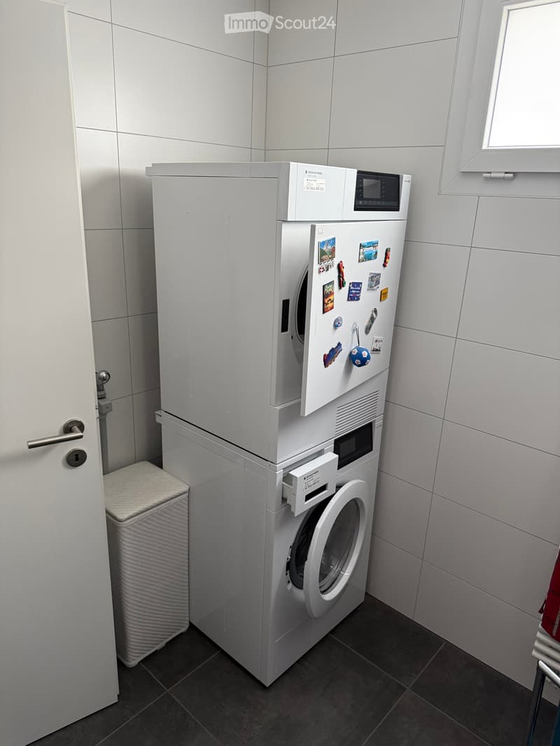 A compact bathroom with a washing machine and a refrigerator, both adorned with various magnets and stickers. The room features white tiled walls and a dark tiled floor.