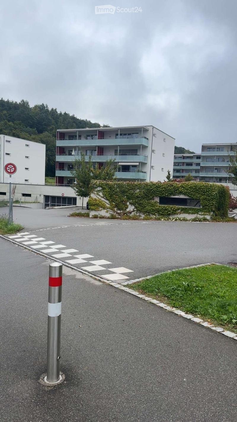 Multi-story residential building with balconies, surrounded by greenery and a paved parking area.