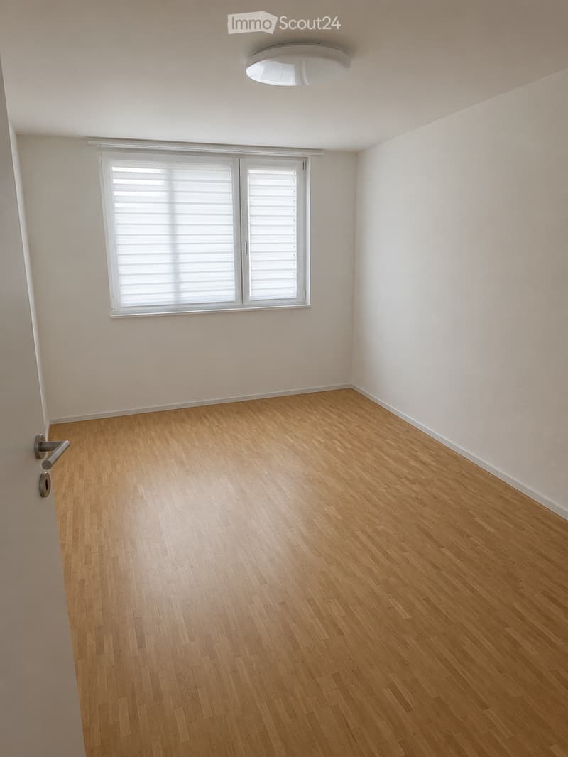 Empty room with light wooden flooring, white walls, and a large window with blinds. The room has a simple, clean design with a single door visible on the left side.