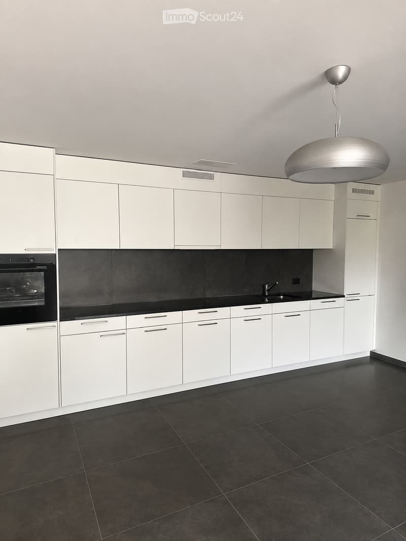 Modern kitchen with white cabinets, black countertops, and a dark tiled floor. Features an integrated oven, sink with faucet, and a sleek pendant light fixture.