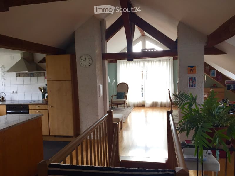 Bright, open living space with wooden floors, exposed wooden beams, and large windows. Features a cozy seating area with a chair and stool, a kitchen area with cabinets and appliances, and a staircase leading to another level.