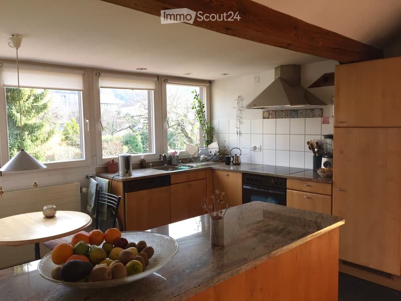 Bright kitchen with wooden cabinets, granite countertop, and large windows. Includes a dining table with a bowl of fruit, a vase with dried flowers, and various appliances like a dishwasher, oven, and blender.