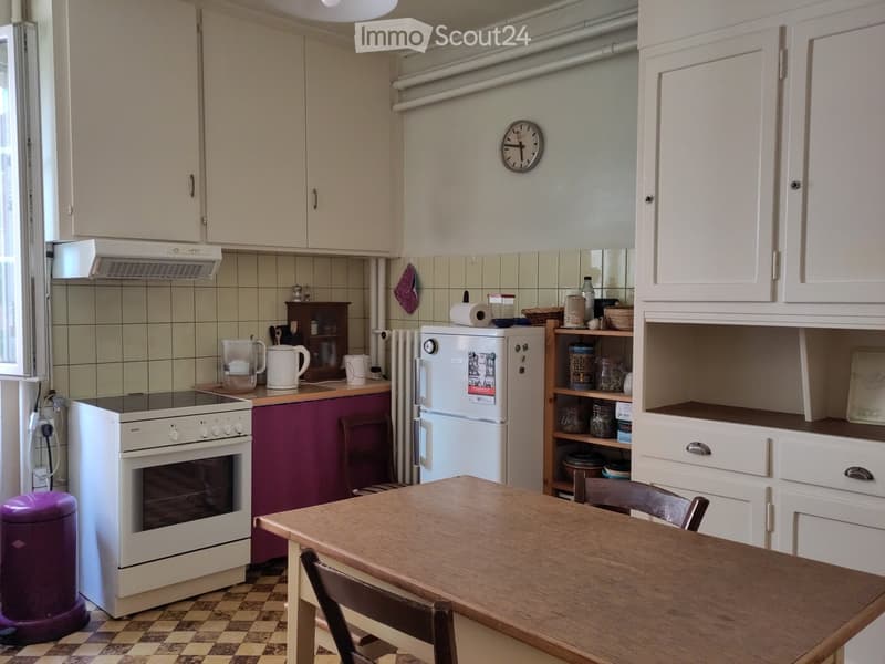 The kitchen features white cabinets, a wooden dining table with chairs, a white oven, a refrigerator, and various kitchen items on the counter and shelves.