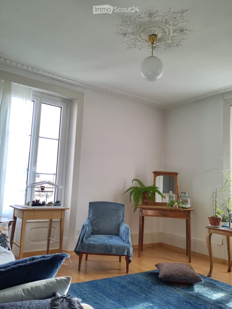 A cozy, well-lit room with wooden flooring, a blue armchair, a small wooden table with a mirror, and several potted plants. The room features a blue rug, a decorative ceiling light, and a window with sheer curtains.
