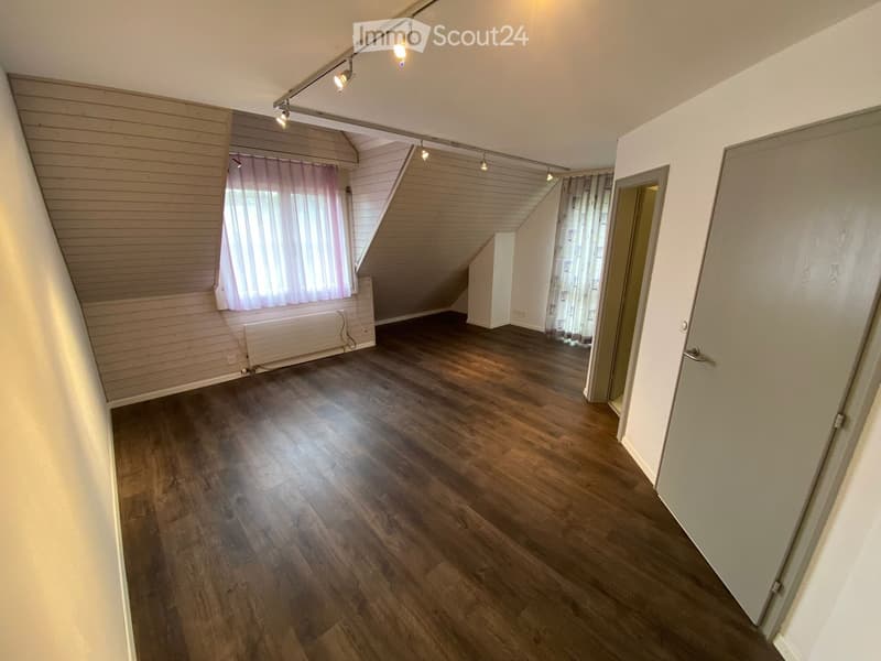 Empty attic room with wooden flooring, slanted ceilings, and large windows with curtains. No furniture or equipment present.