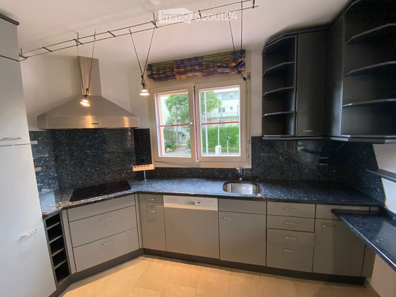 Modern kitchen with grey cabinets, dark granite countertops, stainless steel appliances, and a large window with a view of greenery.