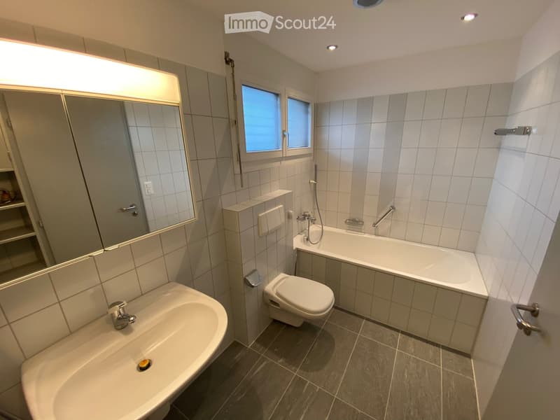 Modern bathroom with a sink, toilet, bathtub, and large mirror. The room features tiled walls and a window providing natural light.