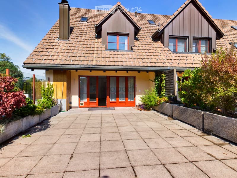 Two-story house with a tiled roof, yellow walls, and red-framed windows. The entrance features large glass doors with curtains, flanked by greenery and a paved walkway.
