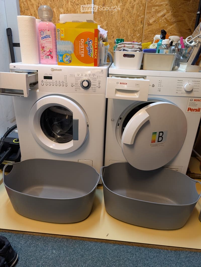 A laundry room with a white washing machine and a white dryer, both Bosch brand, with gray plastic bins placed in front of them, along with storage boxes and cleaning supplies on top of the washing machine