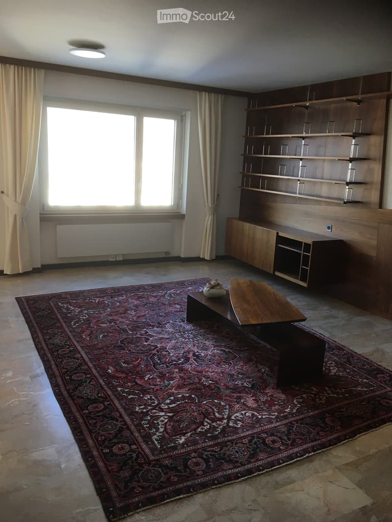 A spacious living room with large windows, wooden shelves, a patterned rug, and a small table.