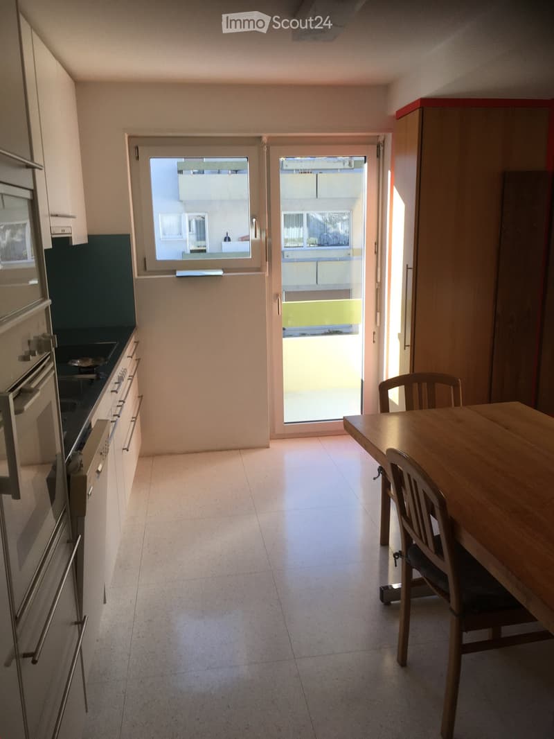 A modern kitchen with white cabinets, a wooden dining table, and chairs, leading to a balcony with a view of the building.