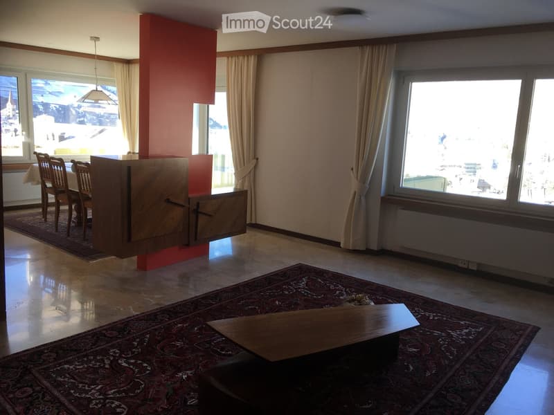Open-plan living and dining area with large windows, wooden furniture, and patterned rugs.