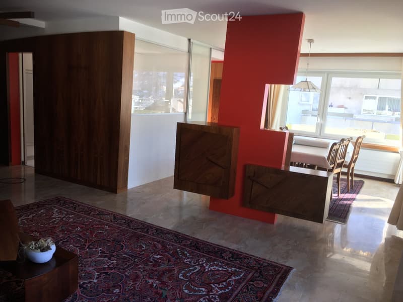 Open-plan living and dining area with large windows, wooden furniture, and a patterned rug.