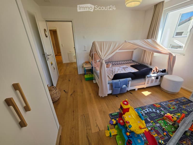 A cozy children's bedroom with a white wooden bed featuring a canopy, a small table with toys, a round white stool, and a colorful play mat with various toys scattered around.