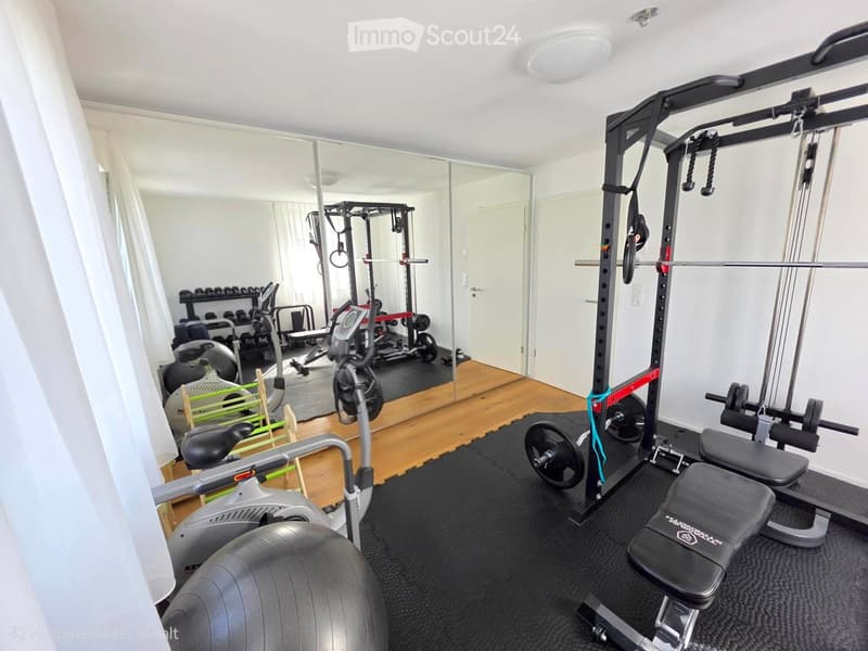 Well-equipped home gym with various exercise equipment including a weight bench, dumbbells, barbell, exercise bike, and a large exercise ball. The space features wooden flooring and large mirrors.
