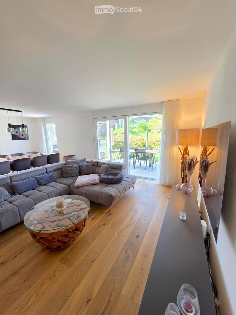 Modern living room with a grey sectional sofa, glass coffee table, dining table with chairs, and decorative lamps. Large sliding glass doors lead to a patio with outdoor furniture.