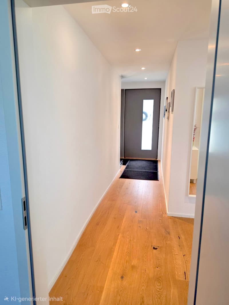 Clean, modern hallway with light wooden flooring, white walls, and recessed ceiling lights. A gray door with a window is visible at the end, and a small black rug lies in front of it.