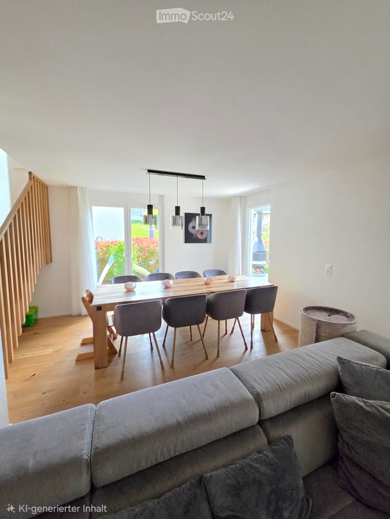Bright living and dining area with a grey sofa, wooden dining table, six chairs, and a cat basket. Large windows and a staircase lead to another level.