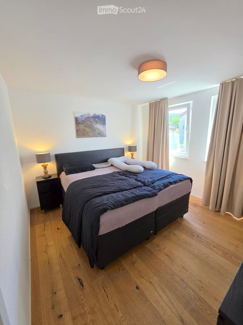 Cozy bedroom with wooden flooring, large bed with dark bedding, nightstand with lamp, and a picture frame on the wall.