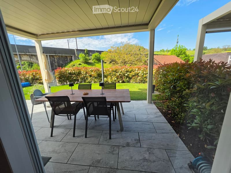 Patio area with a wooden table and chairs, surrounded by greenery and a clear sky.