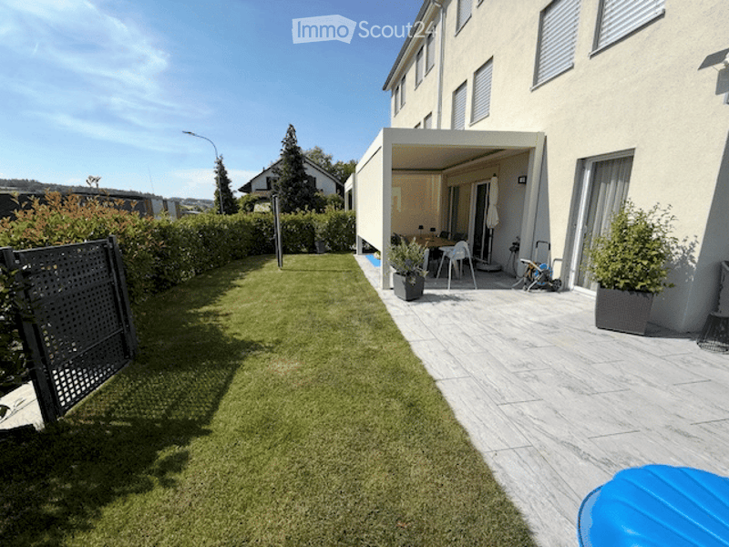 Well-maintained garden with a neatly trimmed lawn, a covered patio area with outdoor furniture, and potted plants. The patio features a table and chairs, and the surrounding area includes a modern house with large windows.