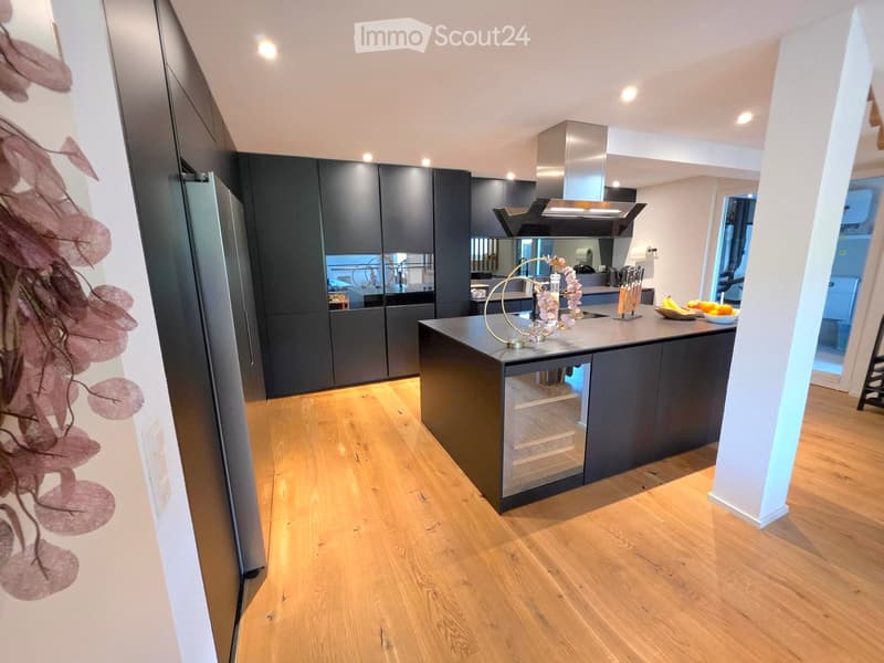 Modern kitchen with dark grey cabinets, a central island, and a stainless steel range hood. The island features a wine cooler and a decorative centerpiece with flowers. The floor is light wooden, and the space is well-lit with recessed lighting.