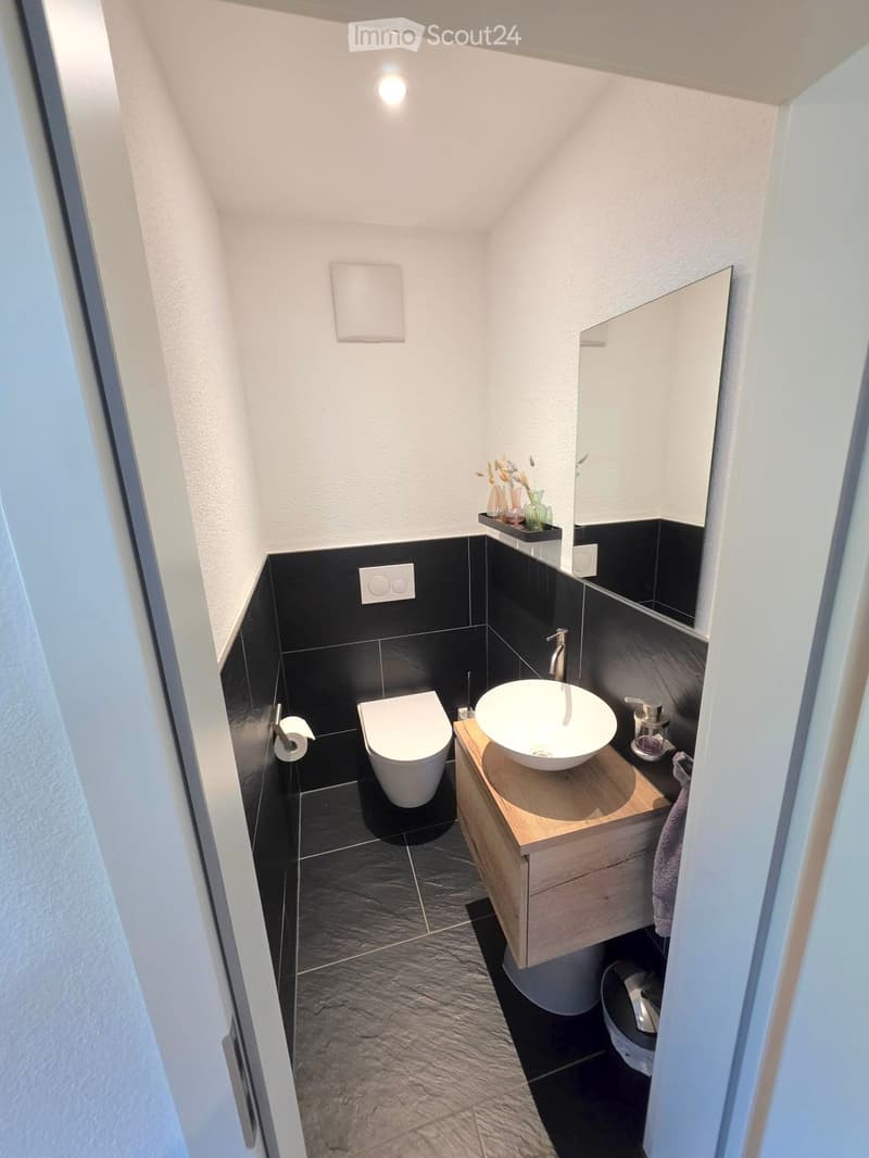 Modern, compact bathroom with black tiled walls and floor, white toilet, wooden sink cabinet, and a round sink bowl.