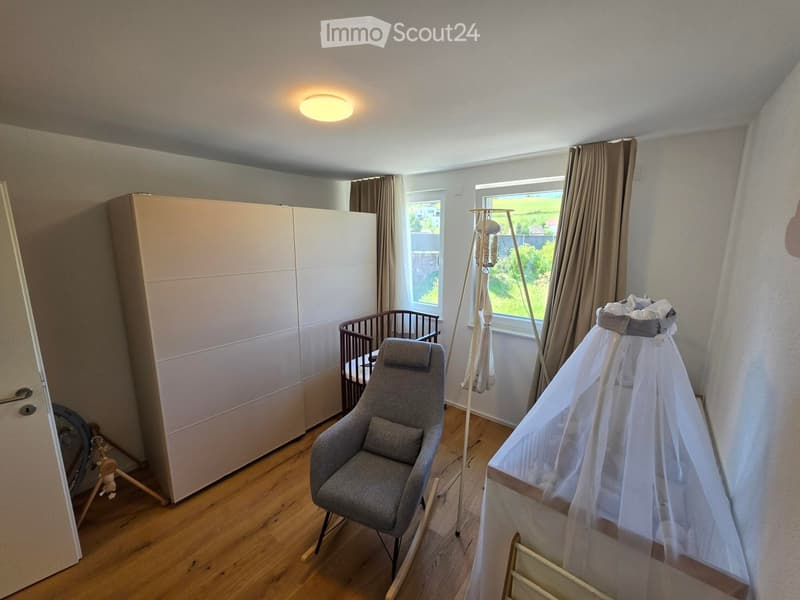 A well-lit nursery with wooden flooring, a grey armchair, a large wardrobe, a baby crib, a baby cot with a canopy, and a standing lamp.