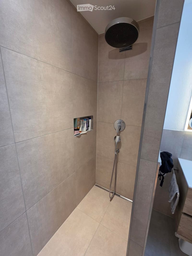 Modern bathroom with beige tiled walls and floor, featuring a handheld showerhead, control valve, and a small shelf holding toiletries. A towel is visible on the side.