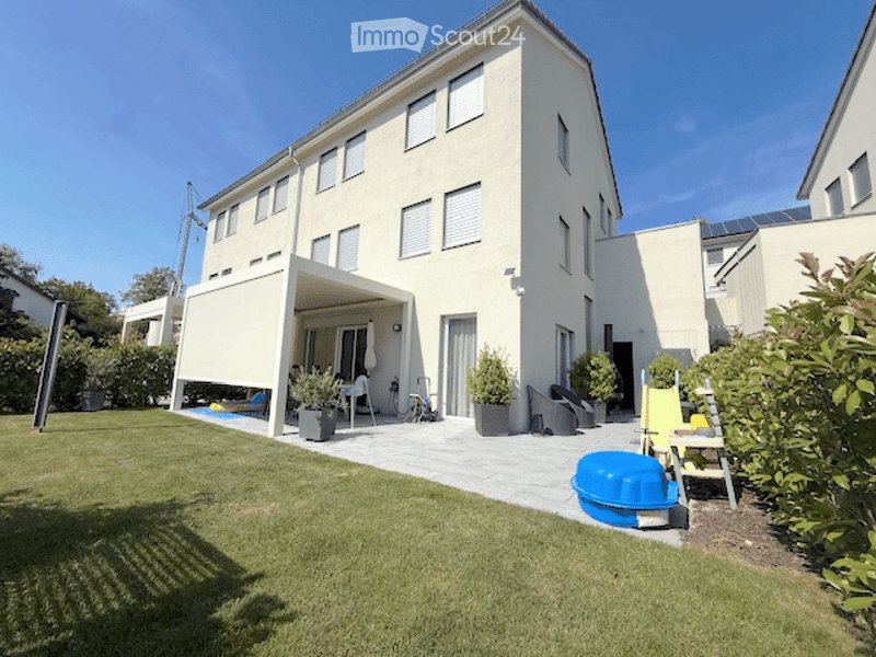 A modern two-story house with a spacious garden and patio area. The patio features a covered seating area with outdoor furniture, potted plants, and a small swimming pool. The lawn is well-maintained, and the house has multiple windows and a clean, contem