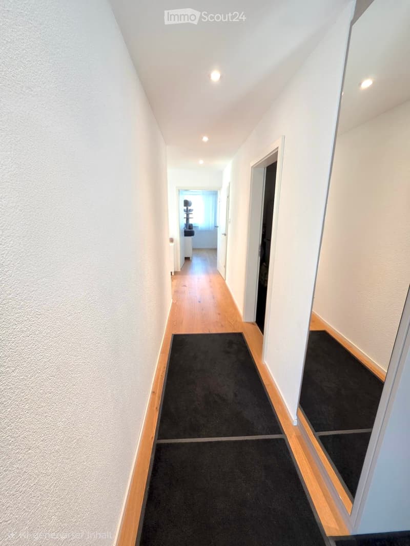 A clean, well-lit hallway with wooden flooring, white walls, and black runner rugs. The ceiling has recessed lighting, and there are several doors leading to other rooms.