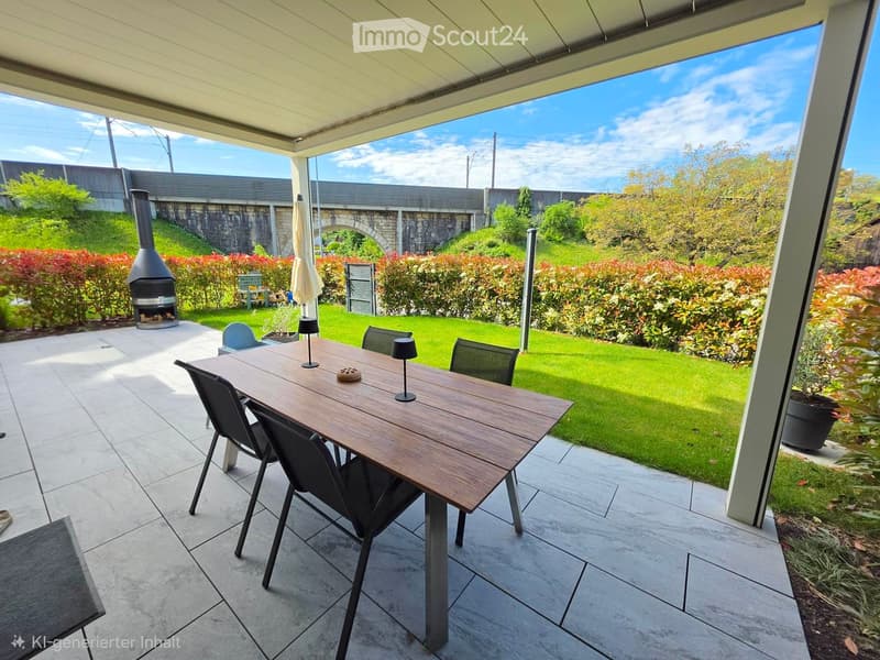 Outdoor patio with a wooden dining table, chairs, and a closed umbrella. A fireplace is visible in the background, along with a well-maintained lawn and garden area.