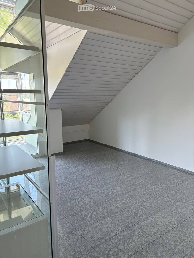 Empty room with a slanted wooden ceiling, tiled floor, and glass balcony railing visible on the left side.