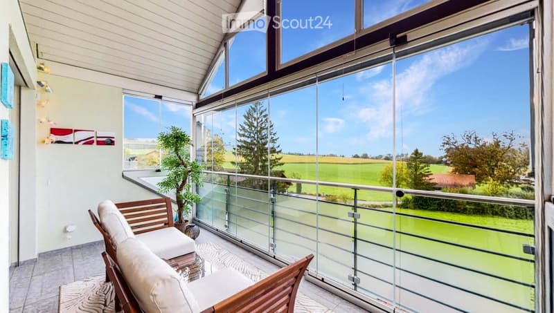 Spacious balcony with large glass windows offering a scenic countryside view, featuring a wooden sofa and a potted plant.