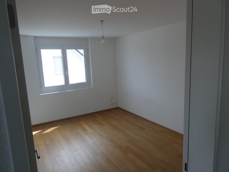 Empty room with light wooden flooring, white walls, and a window with blinds allowing natural light to enter.