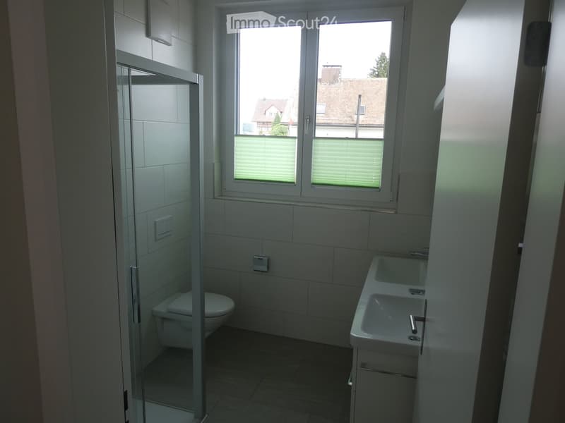 Modern bathroom with a toilet, sink, and glass-enclosed shower area. The room features white tiled walls and a large window with blinds offering a view of the street and neighboring houses.
