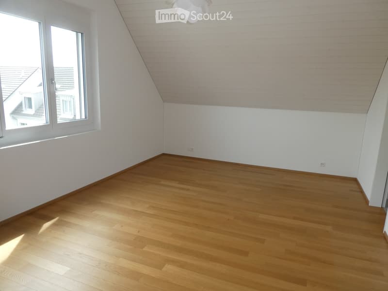 Empty room with wooden flooring, white walls, and a large window letting in natural light. The ceiling has a sloped design typical of an attic space.