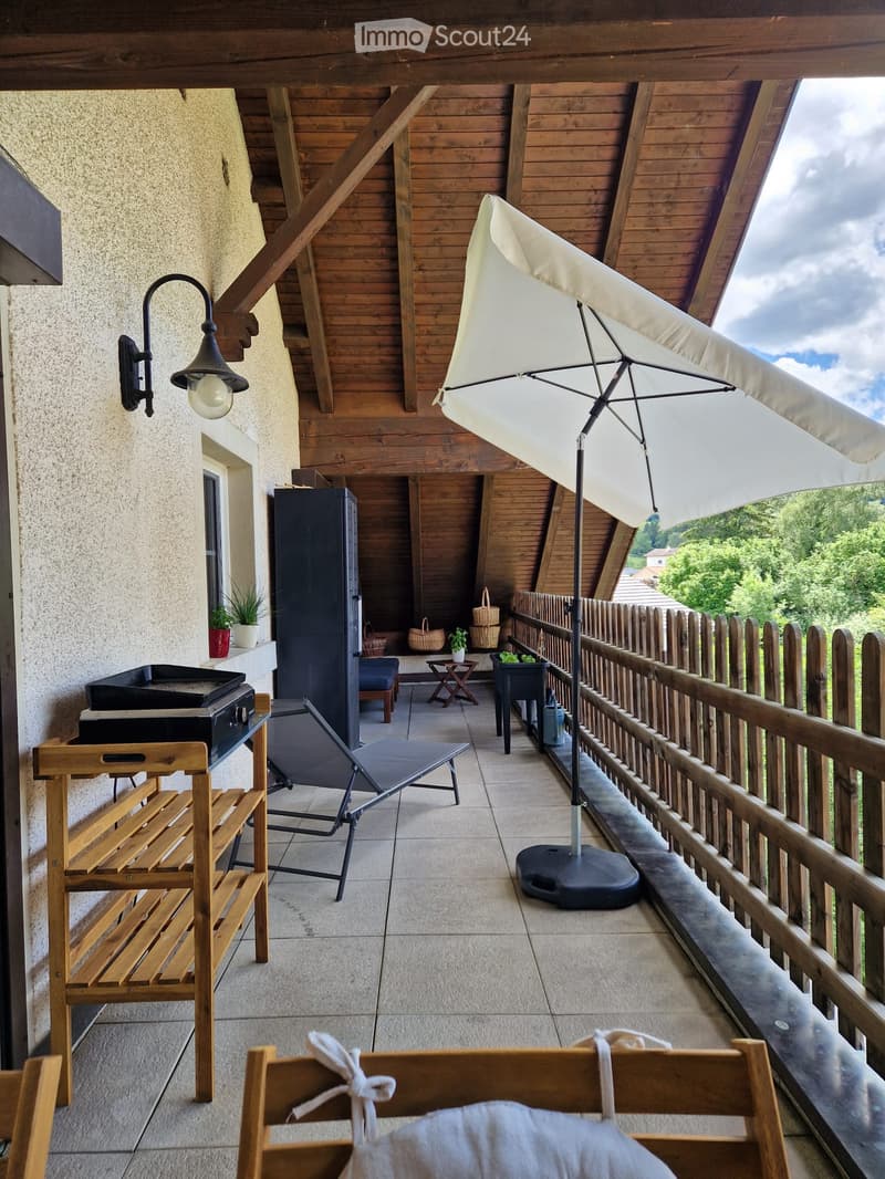 Balcony with wooden beams, tiled floor, umbrella, wooden bench, lounge chair, small table, and potted plants.