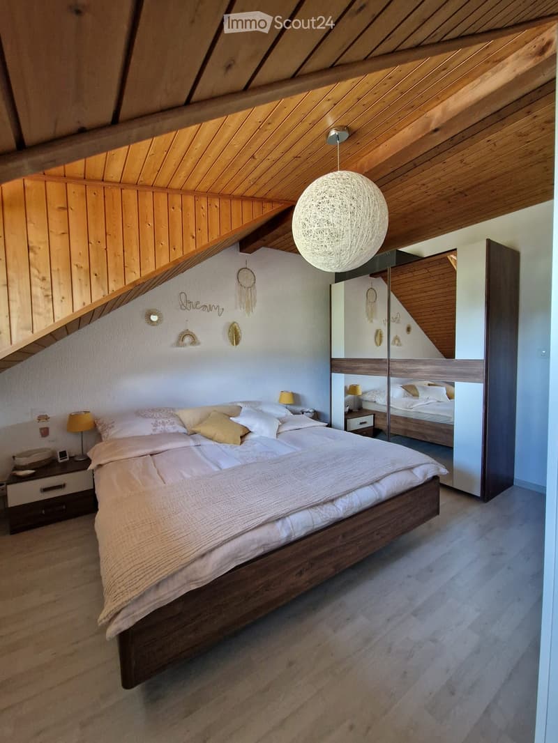 Cozy attic bedroom with wooden slanted ceilings, a large bed with white bedding, nightstands with lamps, and a mirrored closet. Decorative wall art and a pendant light are visible.