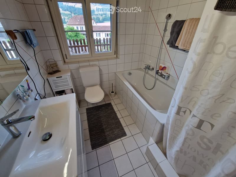 Bathroom with a bathtub, toilet, sink, mirror, and a small cabinet. Towels are placed on the edge of the bathtub, and a window offers a view of the outdoor area.