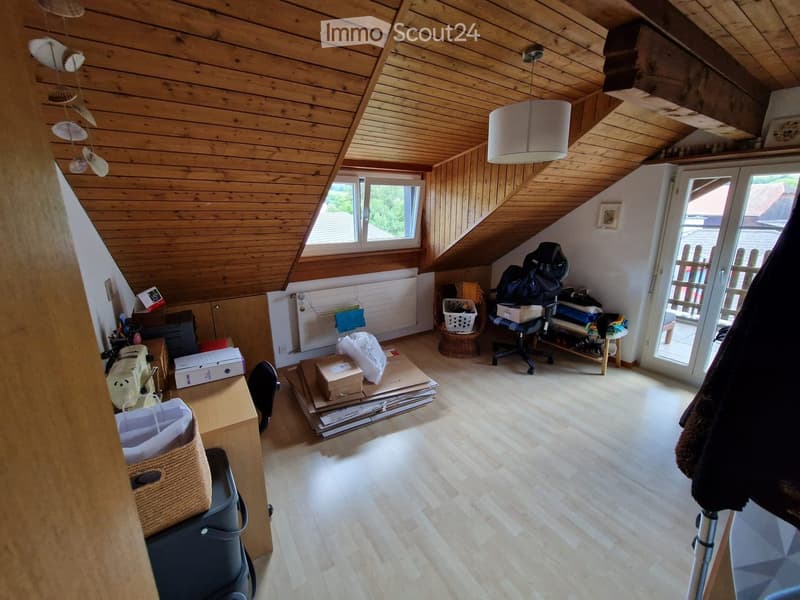 Bright attic room with wooden ceiling and flooring, featuring a desk, chair, wicker basket, and various storage items.