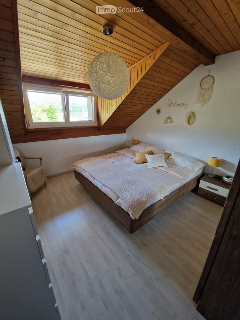 Cozy attic bedroom with wooden slanted ceilings, large windows, a wooden bed with white bedding, a nightstand with a lamp, and a decorative wall hanging.