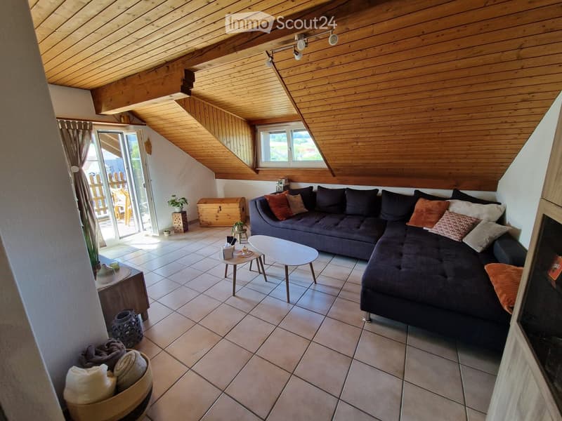 Cozy living room with a dark grey sectional sofa, multiple throw pillows, a white coffee table, side table, and a chest. The room features wooden beams on the ceiling and large windows letting in natural light.