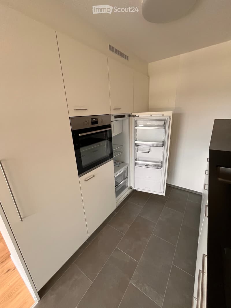 Modern kitchen with light-colored cabinets, dark tiled floor, and stainless steel appliances including an oven and a refrigerator.