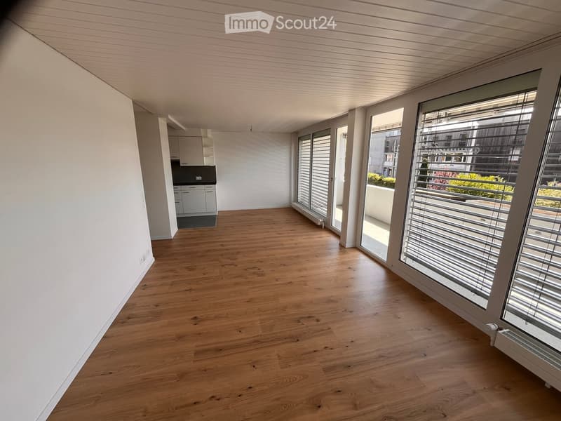 Empty room with wooden flooring, white walls, and large windows with blinds. A small kitchen area is visible in the background with cabinets and appliances.