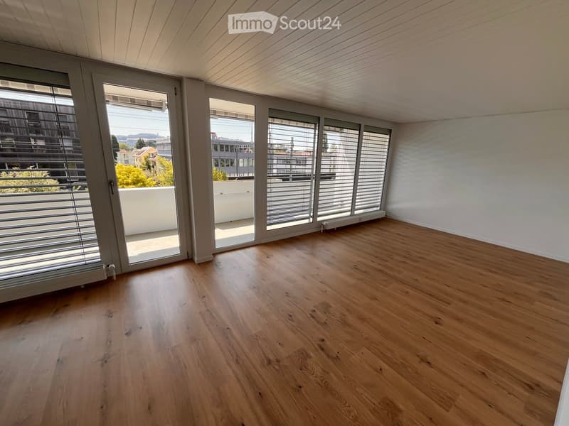 Empty room with wooden flooring, large windows with blinds, and a view of the city and greenery outside.