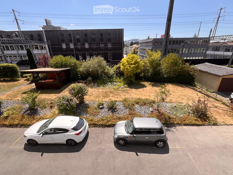 A small garden area with a wooden gazebo, surrounded by bushes and trees, with two parked cars in the foreground.