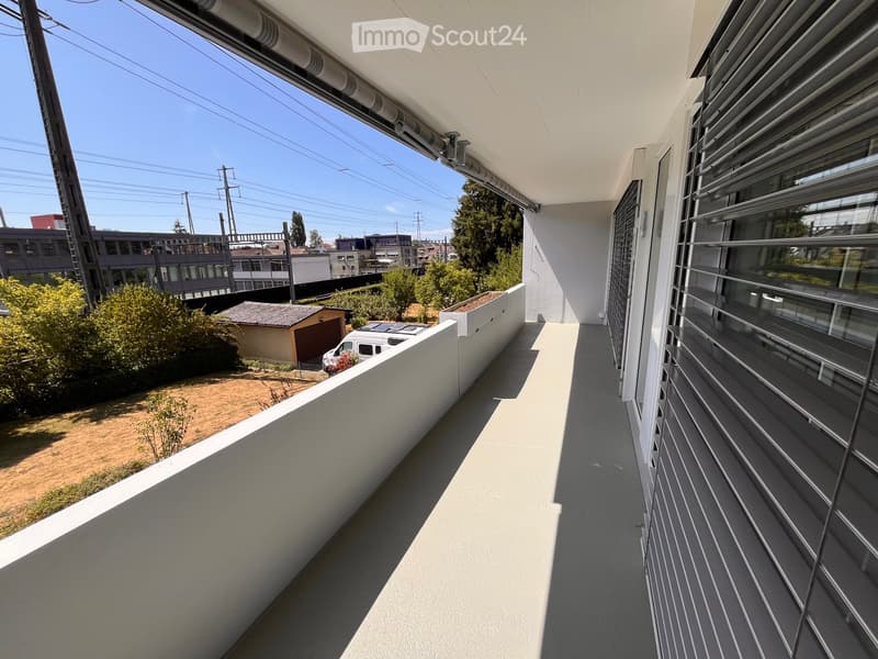 Modern balcony with white walls, grey flooring, metal railings, and a view of the surrounding area including buildings, trees, and a parked van.