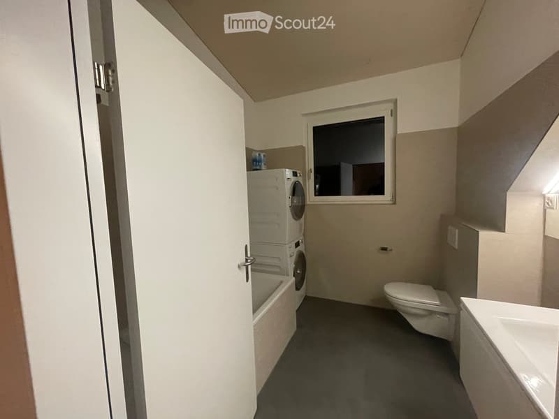 Bathroom with a toilet, sink, bathtub, and a washing machine placed near the window. The room has light-colored walls and a grey floor.