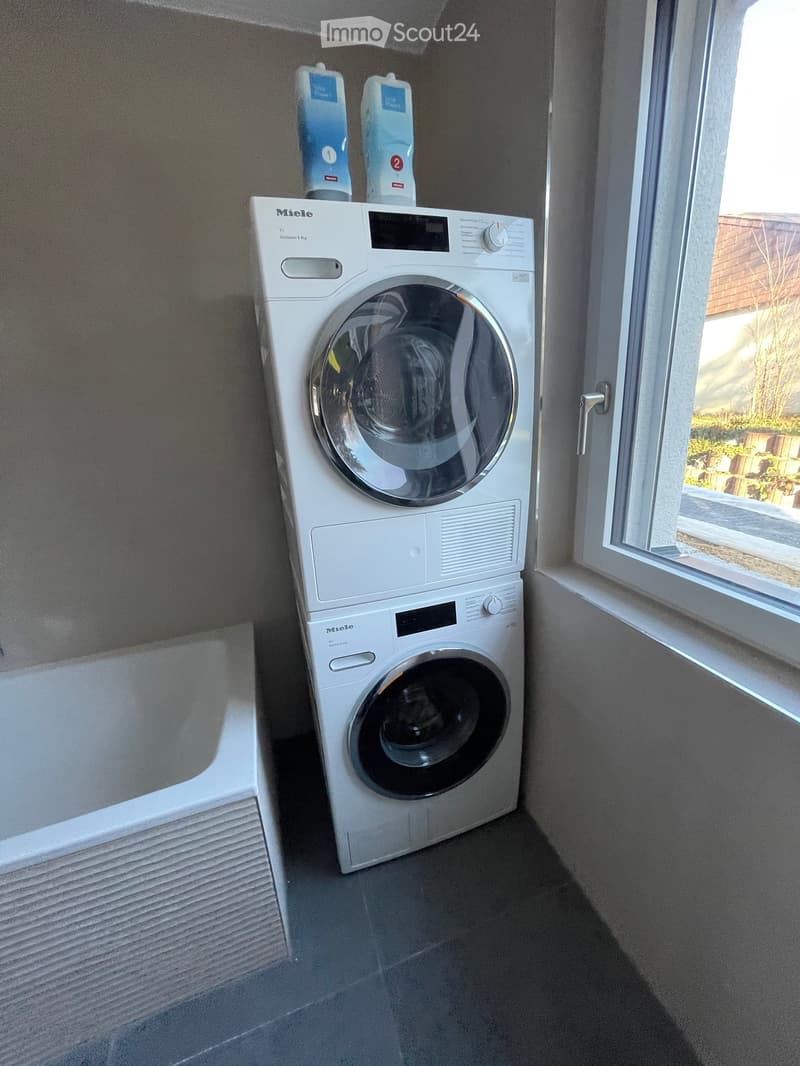A modern bathroom with a stacked washing machine and dryer unit, featuring a sleek white design and digital control panels.